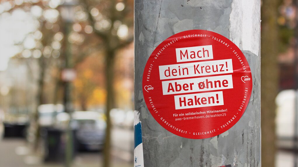 Das Foto zeigt einen Aufkleber, der auf einer Straßenlaterne geklebt wurde. Auf dem Aufkleber steht: Mach Dein Kreuz! Aber ohne Haken